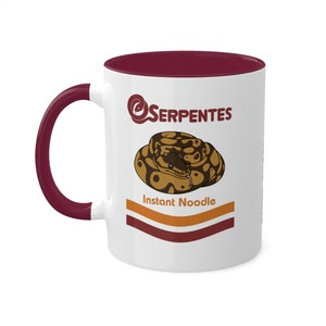 May include: White ceramic mug with a maroon rim and handle. The mug features a cartoon illustration of a snake curled up in a bowl with the text "Serpentes Instant Noodle" printed on the front.