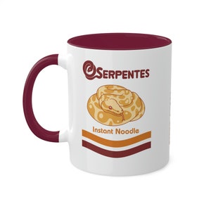 May include: White ceramic mug with a burgundy interior and handle. The mug features a cartoon illustration of a snake coiled in a circle, with the text "Serpentes Instant Noodle" in a playful font.