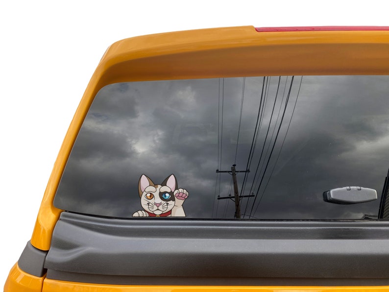 May include: A white, black, and orange cat with one blue eye and one brown eye peeks out of a car window. The cat is wearing a red collar with a gold bell. The car window is tinted and reflects a cloudy sky and power lines.