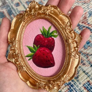 May include: A small, gold-colored, ornate frame holds a painting of two ripe strawberries with green leaves against a pink background. The frame has an oval opening and is held in a hand.