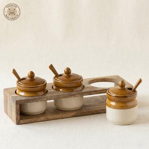 May include: A set of three ceramic condiment jars with brown lids and matching spoons, housed in a rectangular wooden stand. Each jar features a cream-colored base and a glossy brown top. The logo Indian Kalakriti is visible in the top left corner.