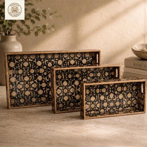 Könnte beinhalten: Set aus drei dekorativen Holztabletts mit floralem Muster. Die Tabletts haben einen schwarzen Hintergrund mit einem goldenen Blumendesign. Die Tabletts sind rechteckig mit Holzrahmen und Griffen. Das größte Tablett ist ca. 46 cm lang.