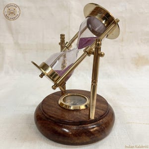 May include: A rotating hourglass with a polished brass frame and clear glass bulbs containing purple sand. The hourglass is mounted on a circular dark brown wooden base that features an integrated brass compass in the center. The logo Indian Kalakriti is visible.