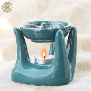 Puede incluir: Un quemador de aceite de cerámica color verde azulado con un cuenco sostenido por tres pilares en forma de mano. Una pequeña vela de té encendida se asienta en la base central, emitiendo humo desde el cuenco. El logo Indian Kalakriti es visible arriba a la izquierda.
