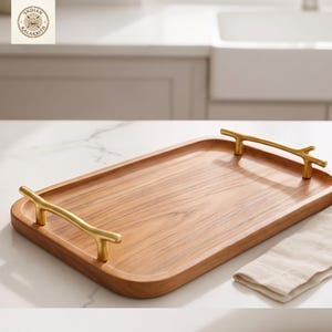 Könnte beinhalten: Ein rechteckiges Serviertablett aus Holz mit hellbraunem Finish. Das Tablett hat goldfarbene, astförmige Griffe an jedem Ende. Auf der rechten Seite des Tabletts liegt eine weiße Stoffserviette.