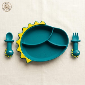 May include: A teal silicone divided plate shaped like a dinosaur with yellow spikes along the edge, accompanied by a matching teal spoon and fork, each featuring a small dinosaur head design at the base of the handle and a yellow spike detail.