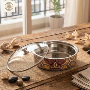 May include: A stainless steel serving bowl with a glass lid. The bowl features a decorative pattern in shades of red, yellow, and brown. The lid has a small, round handle. The bowl sits on a burlap runner on a wooden table.