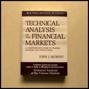May include: A book titled "Technical Analysis of the Financial Markets" by John J. Murphy. The cover is white with brown and beige stripes and text. The book is a comprehensive guide to trading methods and applications.