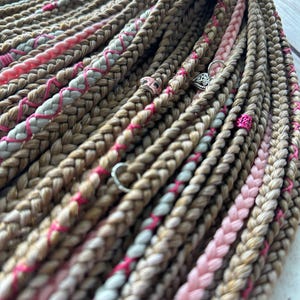 May include: Close-up of braided hair extensions in shades of brown, pink, and white. The braids are adorned with silver-coloured charms and pink accents, creating a textured and colourful look. The hair extensions are arranged in a layered pattern.