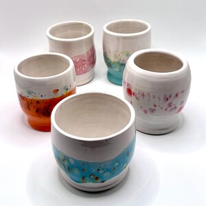 May include: Five handmade ceramic cups with a white base and colorful accents. The cups feature orange, pink, blue, and green speckled designs. The cups are cylindrical with a slightly flared rim, perfect for beverages.