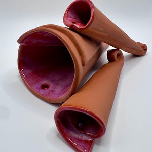 May include: Three terracotta-colored ceramic sculptures with a flared, trumpet-like shape. The interior of each piece is glazed in a vibrant red-purple hue, with a glossy finish. The sculptures are arranged on a white surface, showcasing their unique forms.