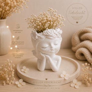 May include: A white ceramic planter shaped like a person's head with closed eyes and a floral crown, filled with dried baby's breath flowers. The planter sits on a round tray, featuring the text ATELIER FEDERLEICHT and HANDGEMACHT MIT HERZ.
