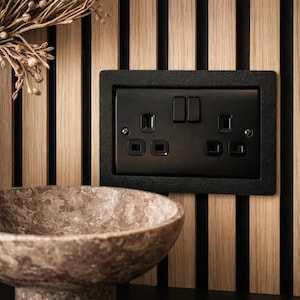 May include: A black double electrical socket with two switches, set into a textured black frame, mounted on a wooden panel with vertical black stripes. A stone bowl is in the foreground. Dried plants are on the left.