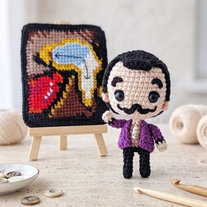 May include: A crochet doll with black hair and a mustache, wearing a purple jacket, standing next to a small wooden easel displaying a square crochet tapestry featuring a yellow melting clock design on a black background with red accents.