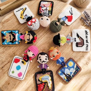 Puede incluir: Una colección de pequeñas muñecas de ganchillo que representan a artistas famosos, cada una acompañada de un lienzo en miniatura de punto de cruz. Las figuras incluyen personajes con rasgos distintivos como bigotes, barbas y pelo colorido.
