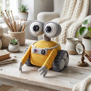 May include: A crochet robot toy featuring a yellow rectangular body, two large round eyes with black and white pupils, and grey articulated arms. The robot has grey treads on the sides and a small control panel on the front with red and black details.