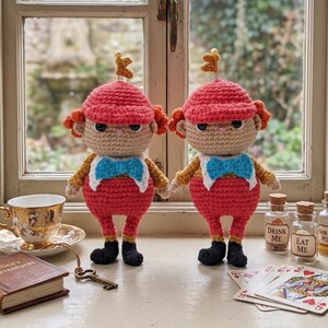 May include: Two identical crochet dolls featuring tan faces, red caps, orange hair, blue bow ties, and red pants with black shoes. Each doll has a small gold key on top of its head. The dolls are displayed with a gold teacup, a book, a key, and two small glass bottles labeled "DRINK ME" and "EAT ME" alongside playing cards.