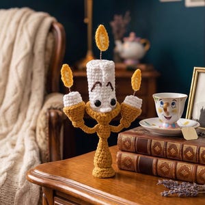 May include: A crochet candelabra figure with a central white candle, two side candles, and three yellow flames. The figure features a gold-colored base and arms, with embroidered facial features including eyes and eyebrows on the central candle.