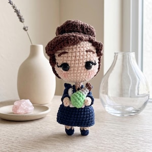 May include: A handcrafted crochet doll featuring a beige face with large black eyes, brown hair styled in an updo, and a dark blue dress. The doll holds a small green and white crocheted potion bottle in its hands, showcasing intricate yarn stitching.