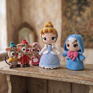 Op de afbeelding: Een verzameling van vijf gehaakte poppen met een centrale figuur in een blauwe jurk met blond haar, een figuur in een blauwe capuchonmantel met een roze strik, en drie kleinere muisachtige figuren in kleurrijke outfits, allemaal met zwarte kraaloogjes.