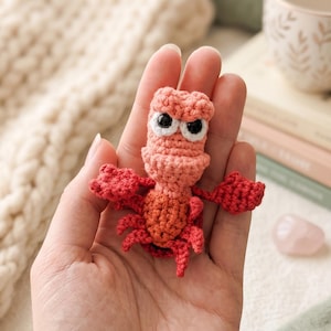 May include: A small, handmade crochet crab figurine featuring a light pink head and a darker red body. The creature has two large, round white eyes with black pupils, two prominent claws extended to the sides, and multiple small legs.