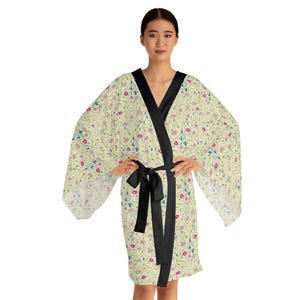 Puede incluir: Una bata kimono color crema con un estampado floral en rosa, azul y verde. La bata tiene un ribete de satén negro y un cinturón negro. Las mangas son anchas y fluidas, y la bata llega a la rodilla.