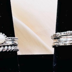 May include: Two silver rings on black stands. One ring features a pearl, the other a green gemstone. The rings have intricate designs, including a square, twisted rope, and floral patterns. The rings are displayed on a light-colored background.