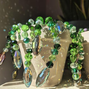 Può includere: Un portapiante decorativo con perline di cristallo verdi e iridescenti. Le perline sono infilate insieme in varie forme e dimensioni, creando un effetto a cascata. Il portapiante è progettato per essere appeso a un gancio o a una struttura simile.