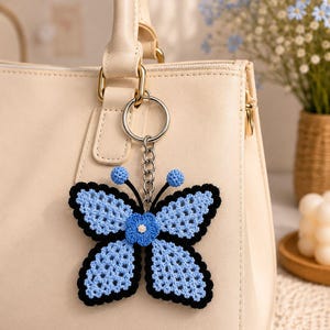 May include: A light blue and black crochet butterfly keychain hanging from a cream-colored handbag. The butterfly has a blue flower in the center and blue pom-poms on the antennae. The handbag has gold hardware.