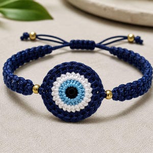 May include: A navy blue crochet bracelet featuring a central evil eye design. The eye is white, light blue, and black. Gold-colored beads accent the bracelet, which is adjustable with navy blue cords.