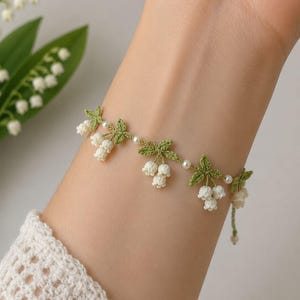 Puede incluir: Un brazalete de ganchillo hecho a mano con un diseño de lirios del valle. El brazalete presenta pequeños racimos de flores blancas, hojas verdes y delicados detalles de perlas. El brazalete se lleva en una muñeca, mostrando los intrincados detalles del patrón floral.