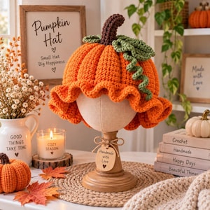 May include: A bright orange crochet pumpkin hat with a ruffled brim, a dark brown stem, and a textured green leaf and vine detail. The hat is displayed on a wooden stand with a tag that reads Handmade with Love, surrounded by autumn decor.