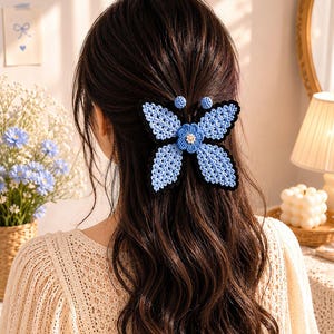May include: A light blue and black crochet butterfly hair clip. The butterfly hair accessory is attached to dark brown hair. The butterfly has a flower in the center and two small blue circles above the butterfly.