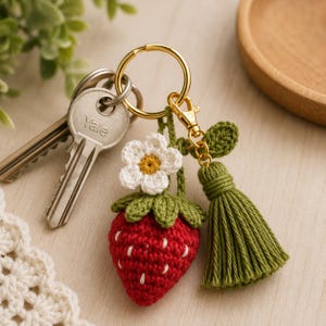 Könnte beinhalten: Ein handgefertigter roter Häkel-Erdbeer-Schlüsselanhänger mit weißen Samendetails, grünen Blättern und einer weißen und gelben Blume. Der Schlüsselanhänger enthält einen goldfarbenen Ring und Verschluss sowie eine grüne Quaste.