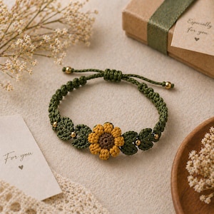 Puede incluir: Una pulsera de macramé verde hecha a mano con un girasol amarillo y marrón y hojas verdes. La pulsera tiene cuentas doradas y un cierre de nudo corredizo ajustable. Una pequeña caja de regalo y una tarjeta que dice "For you" están en el fondo.