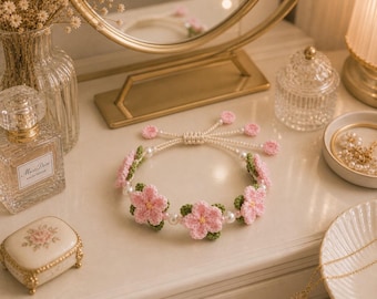 Pode incluir: Uma pulseira de crochê feita à mão com flores rosa e folhas verdes, intercaladas com pérolas brancas. A pulseira está sobre uma superfície branca com outros itens decorativos, incluindo um frasco de perfume e uma pequena caixa de joias.