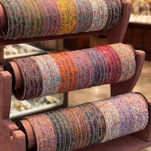 May include: A collection of thin, circular bangles arranged on a three-tiered display stand. Each bangle features intricate, colorful floral patterns and delicate metallic detailing, showcasing a variety of hues including yellow, red, blue, and green.