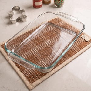 May include: A rectangular clear glass baking dish with integrated handles, resting on a brown and white plaid quilted fabric mat. A metal gingerbread man cookie cutter is positioned to the upper left of the glass dish.