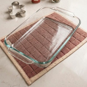 May include: A rectangular clear glass baking dish sits on a quilted fabric mat featuring a small red and white checkered pattern with a beige border. A metal gingerbread man cookie cutter is positioned to the left of the baking dish.
