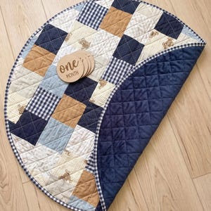 Peut inclure: Un tapis de jeu matelassé circulaire présentant un motif patchwork de carrés bleu marine, beige, bleu clair et crème avec des imprimés d'ours en peluche et des motifs vichy. Un disque en bois avec le texte one MONTH est posé sur le tapis.