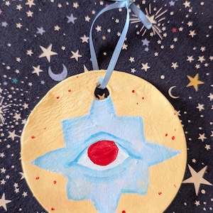 May include: A circular, hand-painted ornament with a pale yellow base and a blue star design. The star has a red eye at its centre. A light blue ribbon is attached for hanging. Small red dots are scattered across the surface.