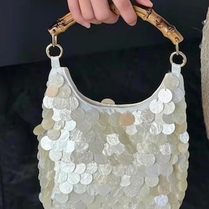 May include: A white handbag covered in overlapping circular iridescent shell-like discs, featuring a curved bamboo handle attached with gold-toned metal rings. The bag has a structured crescent shape and a shimmering, pearlescent surface texture.