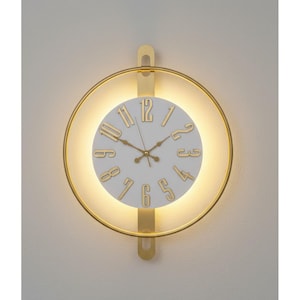 May include: A modern wall clock with a white face and gold-colored numerals and hands. The clock is surrounded by a gold-colored circular frame with a warm, glowing light. The clock is mounted on a light-colored wall.