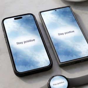Op de afbeelding: Twee smartphones en een smartwatch met de tekst "Stay positive" op hun schermen. De telefoonhoesjes zijn zwart en grijs. Een witte koffiekop en schotel zijn op de achtergrond.
