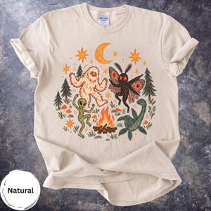 Op de afbeelding: Een naturelkleurig t-shirt met een grafische illustratie van een Bigfoot, een alien, een mothman en het monster van Loch Ness die rond een kampvuur dansen. Het ontwerp bevat oranje sterren, een halve maan, dennenbomen en kleine rode paddenstoelen.