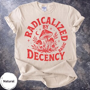 Puede incluir: Una camiseta de manga corta de color natural con un estampado gráfico rojo de setas y follaje. El texto RADICALIZED BY DECENCY está arqueado alrededor de la ilustración central de setas, que incluye pequeñas estrellas y una luna.
