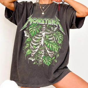 Könnte beinhalten: Ein anthrazitfarbenes Kurzarm-T-Shirt mit einem Grafikdruck eines menschlichen Brustkorbs, der mit grünen Monstera-Blättern und weißen Motten durchsetzt ist. Das Wort MONSTERA ist in einer stilisierten grünen Schrift oben auf dem Design gedruckt.