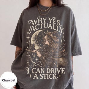 Könnte beinhalten: Ein anthrazitfarbenes Kurzarm-T-Shirt mit einem Grafikdruck einer Hexe auf einem Besen, einem Raben, einer Mondsichel und Sternen. Der Text lautet: WHY YES, ACTUALLY. I CAN DRIVE A STICK. Das Design ist in Creme- und Hellbrauntönen gehalten.