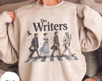 Könnte beinhalten: Ein sandfarbenes Sweatshirt mit Rundhalsausschnitt und einem Grafikdruck von vier historischen Persönlichkeiten, die über einen Zebrastreifen gehen. Über den Figuren steht der Text "The Writers" in einer fetten, dunklen Schrift.