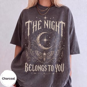 May include: A charcoal gray short-sleeve t-shirt featuring a cream-colored graphic print. The design includes a crescent moon, a moth with a skull pattern, and the text THE NIGHT BELONGS TO YOU in a gothic font, surrounded by ornate floral vines.
